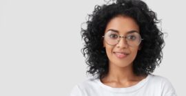 Close up portrait of curly female adult has charming smile, curly dark hair, wears big glasses, satisfied as finished domestic work earlier, being successful designer or architect, has talent